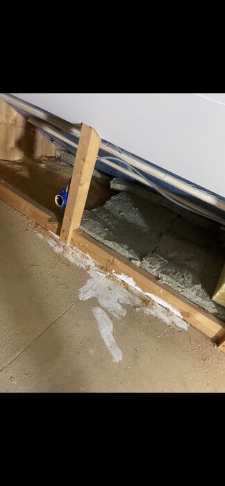 Isolering i kattvind med masonitskivor, träreglar och plast synliga vid byggprojekt, samt blå kablar och ventilationshål med insektsnät i hörnet.