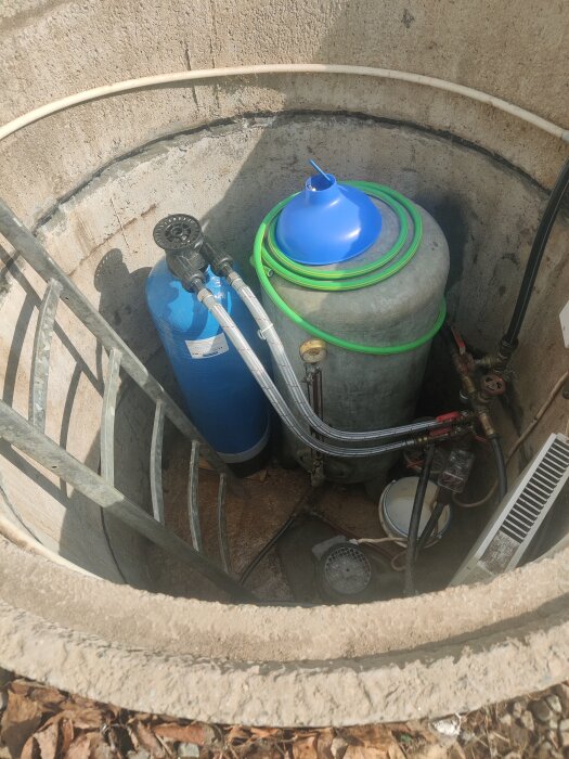 En blå hydrofor och en grön ejektorpump i en betongbrunn, omgiven av slangar och rörledningar med en stege i vänsterkant.