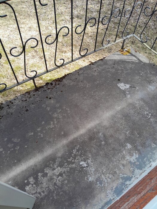 Balkonggolv i betong med flagnande yta och rostfärgad beläggning, beläget bakom ett smidesräcke.