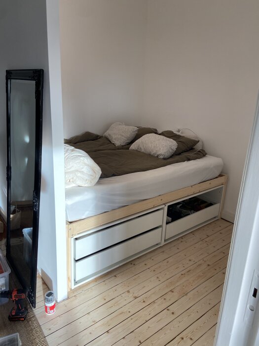 Säng med trälådor i ljus träinramning, vit och olivgrön sängklädsel, bredvid står en svart spegel lutad mot väggen.