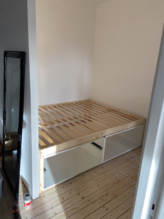 Sängram av trä med vit stomme under, placerad i hörn av rum med trägolv. Spegel och målarburk syns på sidan.