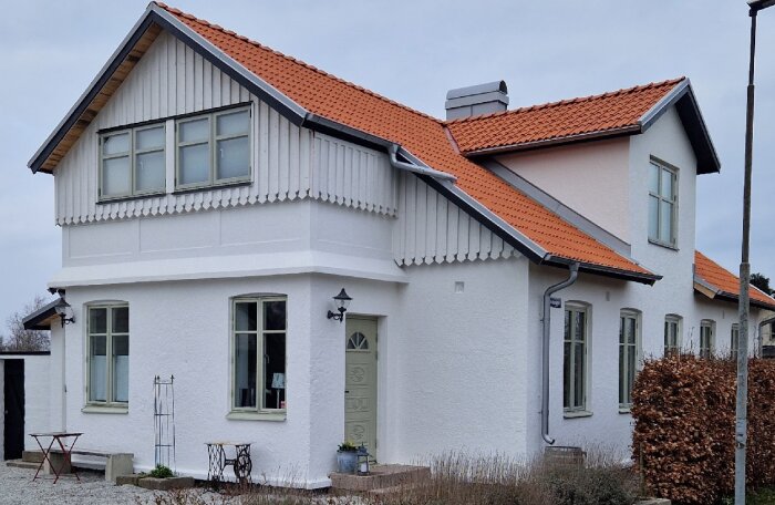 Vitt hus med rött tegeltak, ljusgröna fönster och detaljer vid entrén. Trädgårdsmöbler och buskar i förgrunden.