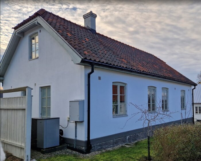 Ett vitputsat hus från 1929 med rött tegeltak och flera fönster längs sidan. En buske och luftvärmepump syns i förgrunden.