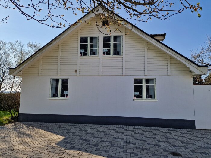 Vitmålad gavel på hus från 1929 med vita spröjsade fönster och svart plåt i solsken. Träd och gräsmatta skymtar i bakgrunden.