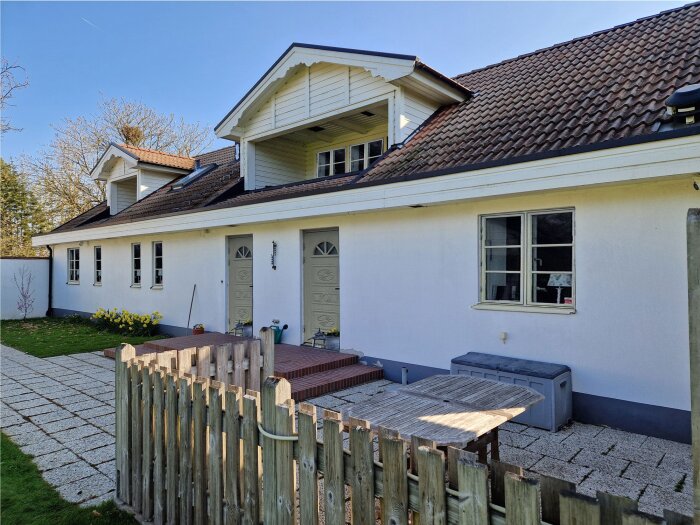 Framsidan av ett hus med vita väggar, två dörrar, och fönster utan spröjs. Trädgårdsstaket och stensatt gång syns i förgrunden.