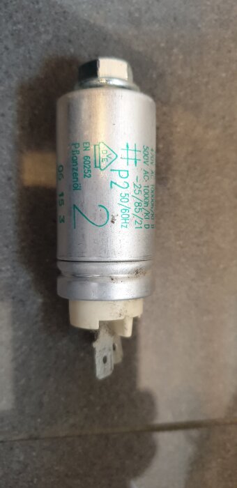 Metallisk plastkondensator med skrift "P2", ansluttande stift synliga, placerad på grå yta, använd i diskussion om köksfläktanslutning.