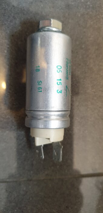 Metallisk kondensator med elektriska anslutningar visas närbild på en grå yta, troligen för en köksfläkt.