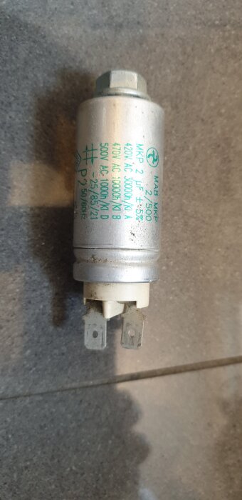Plastkondensator med märkningen MKP 2 μF/500V liggande på en grå yta, använd i diskussion om köksfläktinstallation.