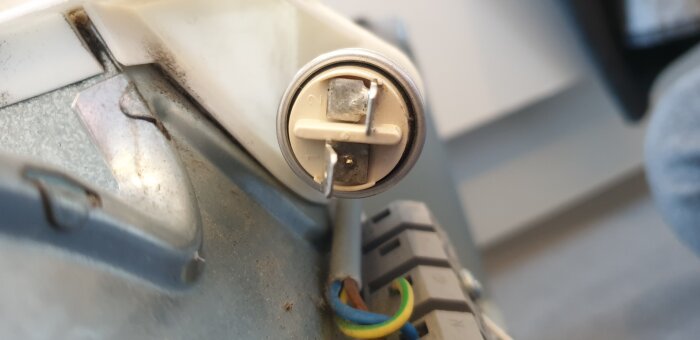 Närbild av elektrisk anslutning på en plastkondensator i en köksfläkt, med synliga stift märkta 1 och 2. Kabeldragning med färger vit, blå och grön/gul.