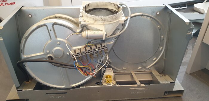 Köksfläktens insida med synliga kablar och plastkondensator, där fläktmotorn är bortkopplad. Kabelfärger: vit, brun och grå.