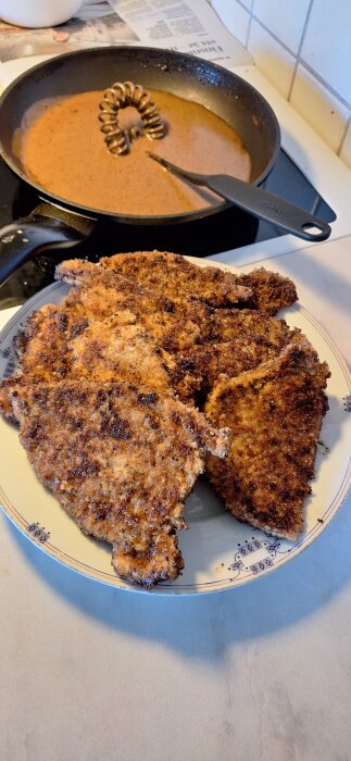 Panerade schnitzlar på en tallrik med en stekpanna med brun sås i bakgrunden.