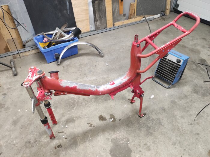 Röd metallram av moped i garageverkstad, med verktyg och tillbehör runtomkring.