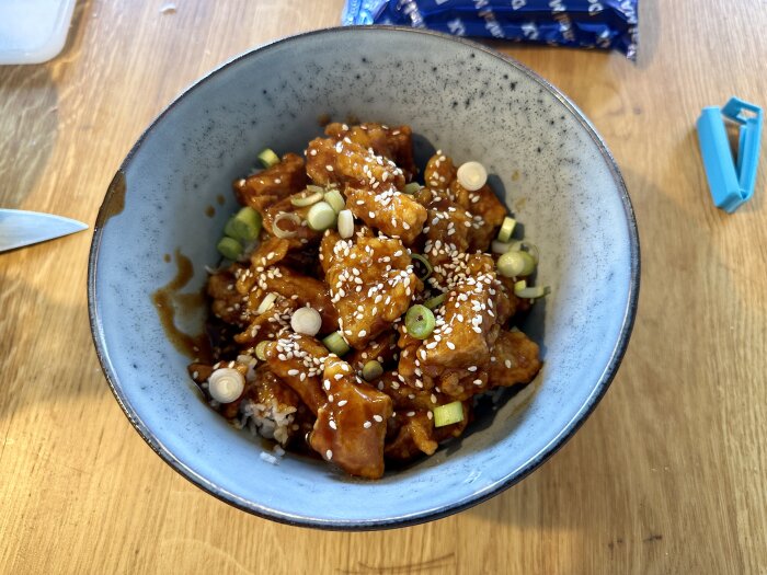 En blå skål fylld med orangefärgade grönsaksbitar, toppade med sesamfrön och skivad grön lök, står på ett träbord.
