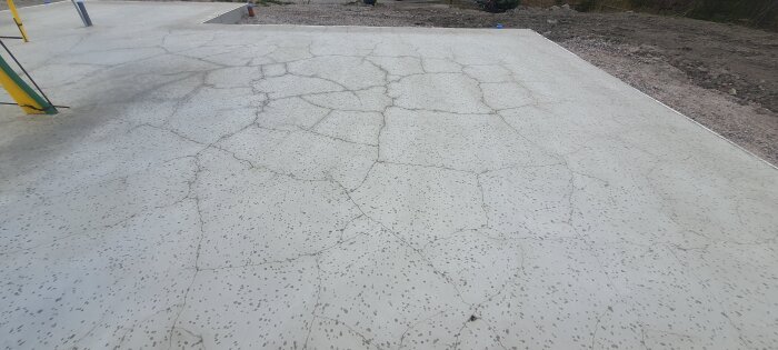 Betongplatta med sprickor över hela ytan, synliga särskilt vid genomföringar. Omgiven av jord och grus, utan synlig efterbehandling.