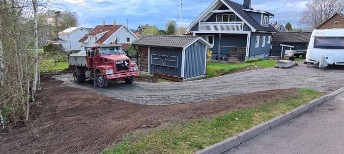 Röd lastbil parkerad på en gårdsplan bredvid ett blått hus, omgiven av grus och pågående markarbete.
