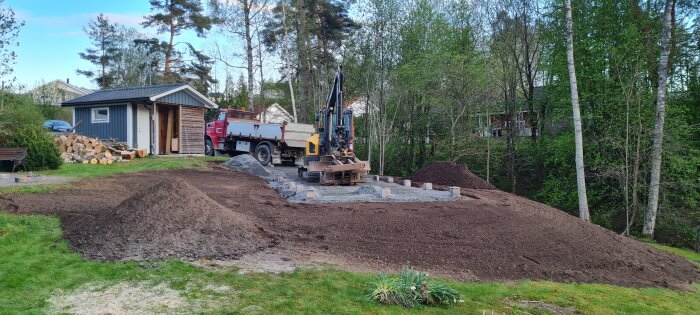 Grävmaskin och lastbil vid friggebod i trädgård, mark förbereds med återvunna kantstenar och grus för nytt beläggningsarbete.