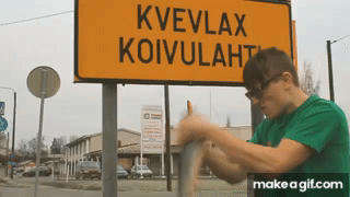 Man i grönt skjorta vevar runt med en fisk framför en skylt med texten "Kvevlax Koivulahti", en humoristisk scen.