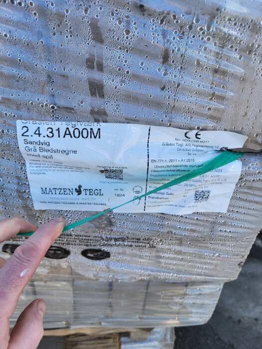 Förpackning med tegel märkt "Grå Blødstrøgne med spil" från Matzen Tegl, täckt av plast med vattendroppar.