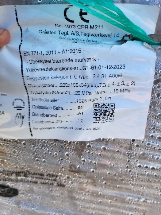 Bilden visar en etikett med bygginformation och specifikationer för murverk, inklusive dimensioner och tryckstyrka, täckt av plast med kondensdroppar.
