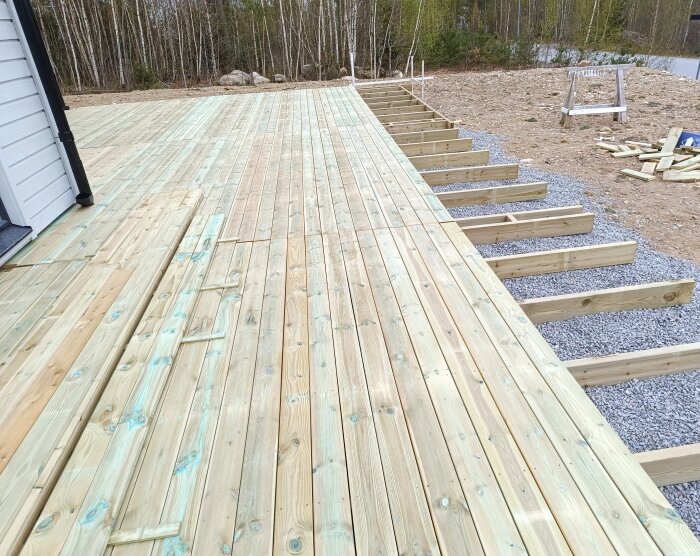 Trädäck under konstruktion, med reglar på grusfundament, markduk och trall som läggs vid sidan av ett hus, omgiven av skog.