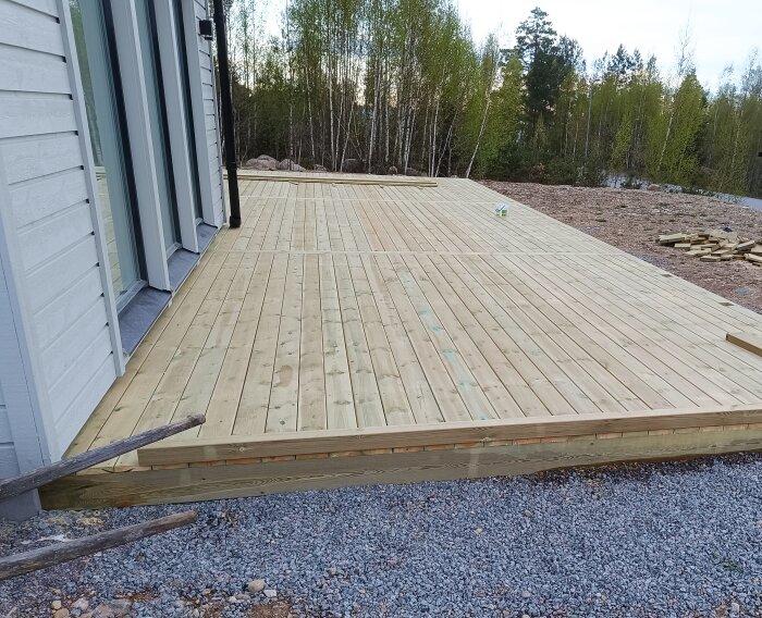 Nybyggt trädäck framför ett hus, med markduk, grus och reglar. Nära skogsområde, brädor synliga på marken.
