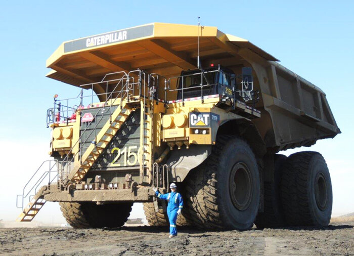 Stor Caterpillar dumper med en person i blå overall framför, jämför storleksmässigt mot en fyrhjuling.