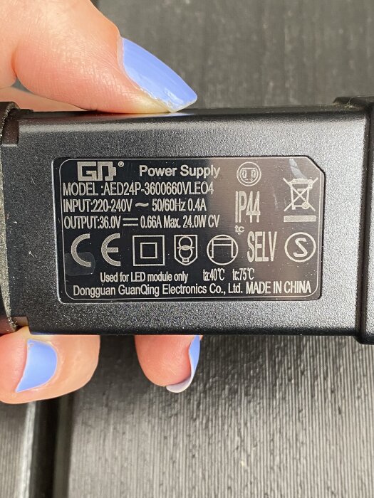 Transformator med etikett, modell AED24P-3600660VLE04, för LED-moduler. Input: 220-240V, Output: 36.0V. Hand med blått nagellack håller transformatorn.