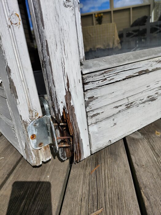 En gammal trädörr med avflagnad färg och skadad trä runt gångjärnet på ett altangolv.