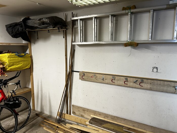 Garagevägg med förvaringshylla, stege, cykel och upphängda trästavar. Tydliga tecken på fuktproblem syns på väggarna.