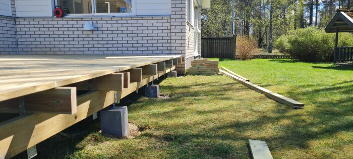 Träveranda under konstruktion med synliga bjälkar och cementpelare, brädor ligger på gräset bredvid. Brickhus i bakgrunden.