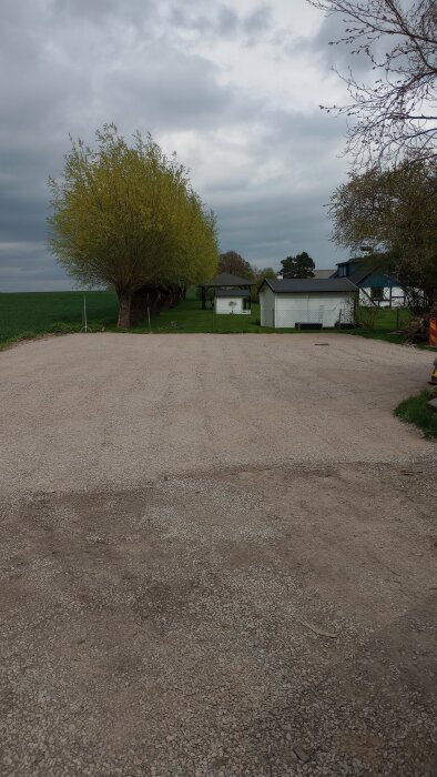 Bilden visar en nyutlagd yta med bärlager framför en grönskande trädgård och bostadshus i bakgrunden.