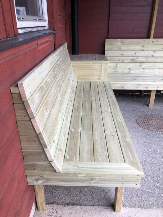 Träbänk med lutande ryggstöd, byggd för kyrkans personal, av obehandlade plankor och placerad mot en röd trävägg.