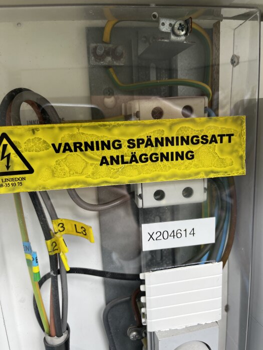 Bild på elcentral med varningsskylt, text 'Varning spänningssatt anläggning', samt kablar märkta L2 och L3.