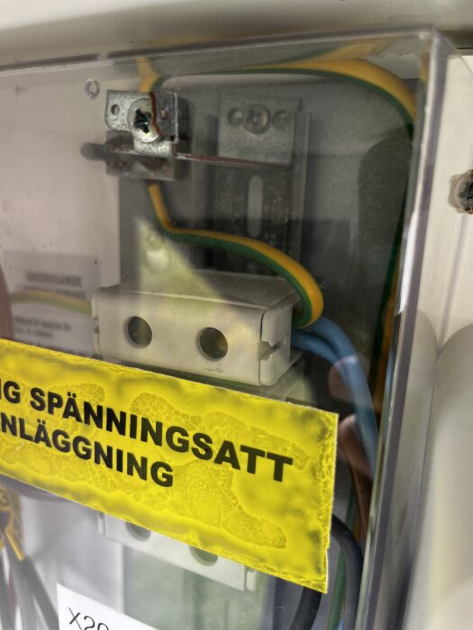 Elcentral med synliga gula och gröna kablar, skruvterminal och gul varningsskylt om spänningssatt anläggning.