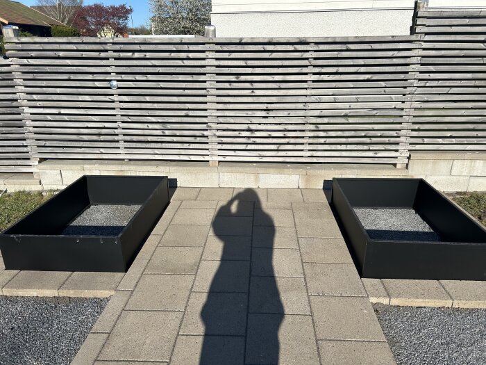 Två svarta pallkragar placerade på en stenlagd yta med grus inuti, framför ett trästaket i en trädgårdsmiljö.
