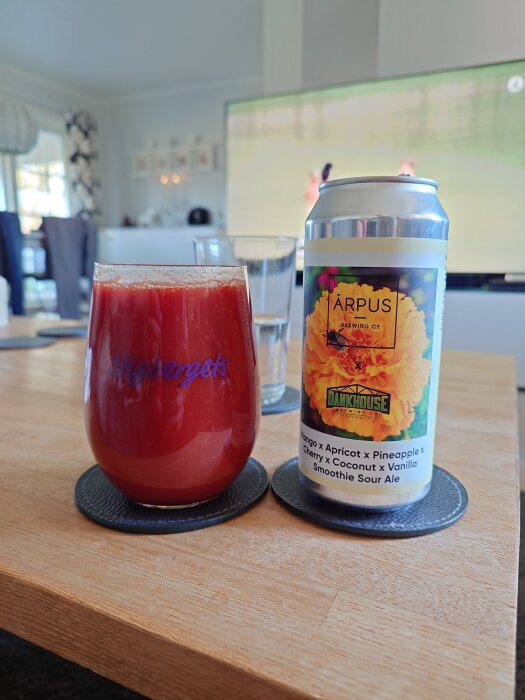 En röd dryck i ett glas bredvid en burk Arpus x Dankhouse smoothie sour ale med fruktig etikett på ett soffbord.