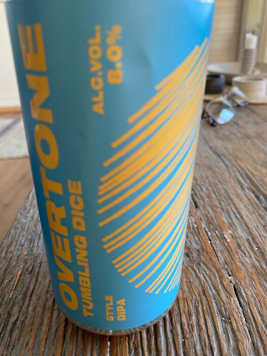 En burk Overtone Tumbling Dice DIPA, 8% alkoholhalt, med blå och gul design stående på ett träbord.