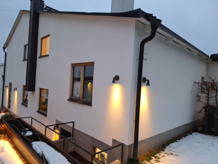 En nyputsad vit fasad med kc bruk, två våningar, svarta stuprör och fönster med trädetaljer, omgiven av snötäckt mark.