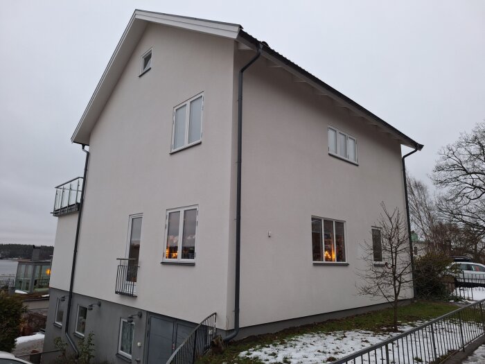 Ett nyputsat hus med vita väggar och svarta stuprör, sett från sidan. Snö på marken och trädgård med få träd.