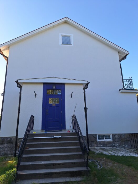 Vit putsad fasad på hus med blå dörr och trappsteg, blå himmel i bakgrunden.