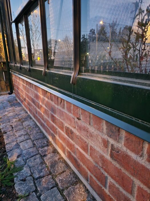 Rödtegelvägg och fönster i kvällsljus med reflektioner och skymtande växtlighet i bakgrunden.