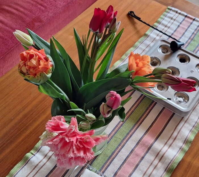 Bukett med färgglada tulpaner i vas på ett randigt underlägg på träbord, med bakgrund av en muffinsform.