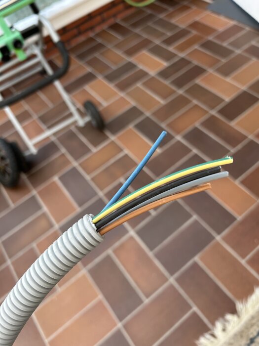 Grå slang med exponerade ledningar i olika färger, liggande på en klinkergolv.