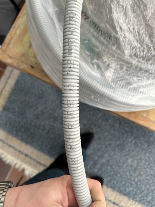 Grå korrugerad slang, NKT Prefix 20mm, hålls av en hand. Rulle med mer slang ses i bakgrunden.