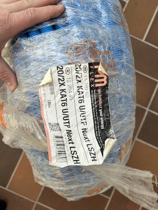 PM Flex 20mm blå slang med dubbel datakabel, 100m. En hand håller i förpackningen med etiketter och produktinformation.