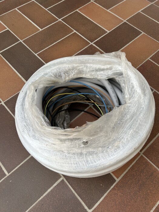 Inplastad rulle med NKT färdigdragen slang och RQ 5G6-kabel, lämplig för elinstallationer, ligger på ett klinkergolv.