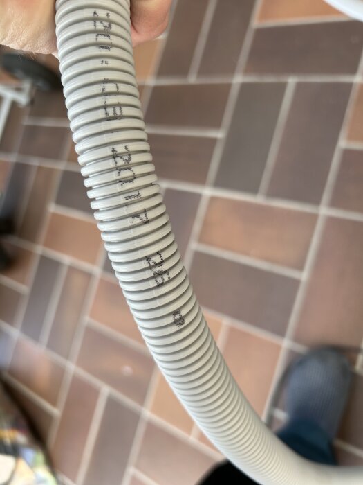 Grå ribbad slang med text, hålls upp på ett stengolv. Används för elinstallation, med modell NKT RQ 5G6.