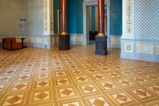 Massivt trägolv med geometriska mönster i ett elegant rum med pelare och dekorerade väggar, typiskt för palats och prestigebyggnader.