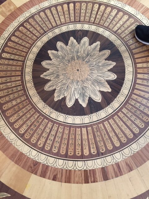 Intrikat trägolvsmönster med cirkulära arrangemang i olika träslag och en central blommig design, liknande golv i palats och slott.