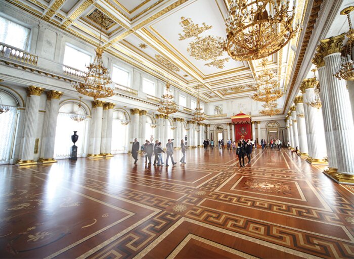 Elegant sal med dekorativa trägolv, utsmyckade tak och stora kristallkronor. Kolonner omger rummet, och besökare går över golvet.